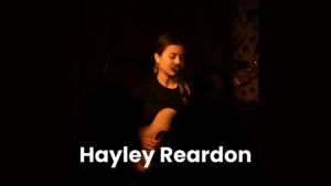Woman holding guitar and singing into microphone with black background.